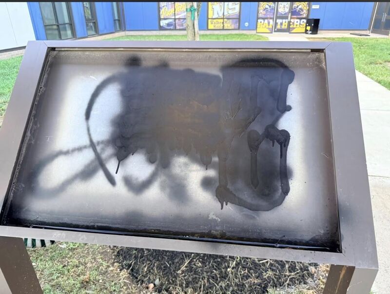 Outdoor school sign display panel completely covered in black spray paint with heavy drips