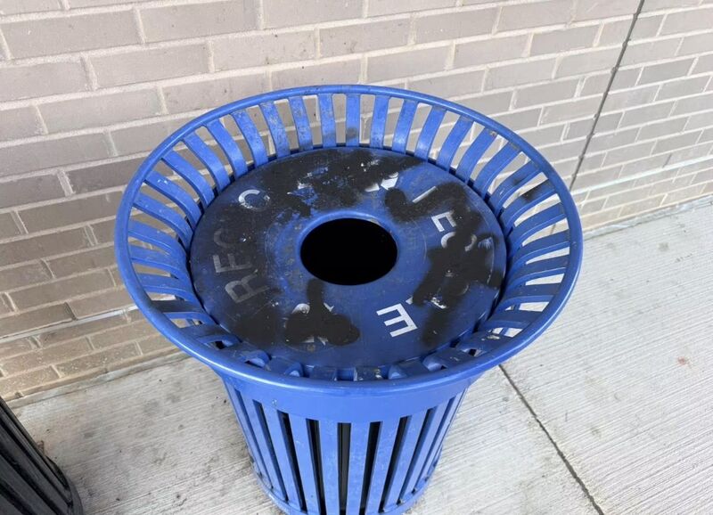Blue metal recycling receptacle with its lid covered in dark spray paint, against a tan brick wall